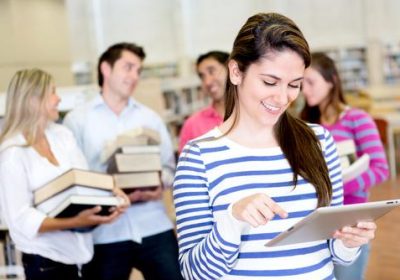 A woman stands with an iPad while three people stand behind her with large books in a library